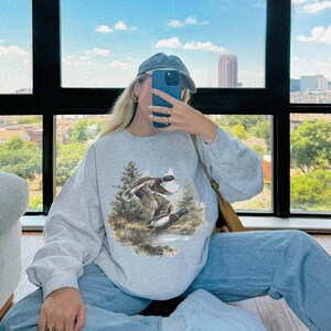 May include: A light gray sweatshirt featuring a detailed illustration of two ducks in flight over a body of water, surrounded by trees. The sweatshirt is paired with light blue jeans, a blue cap, and white sneakers. The word "ASH" is visible in the bottom right corner.