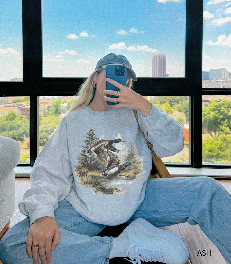 May include: A light gray sweatshirt featuring a detailed illustration of two ducks in flight over a body of water, surrounded by trees. The sweatshirt is paired with light blue jeans, a blue cap, and white sneakers. The word "ASH" is visible in the bottom right corner.