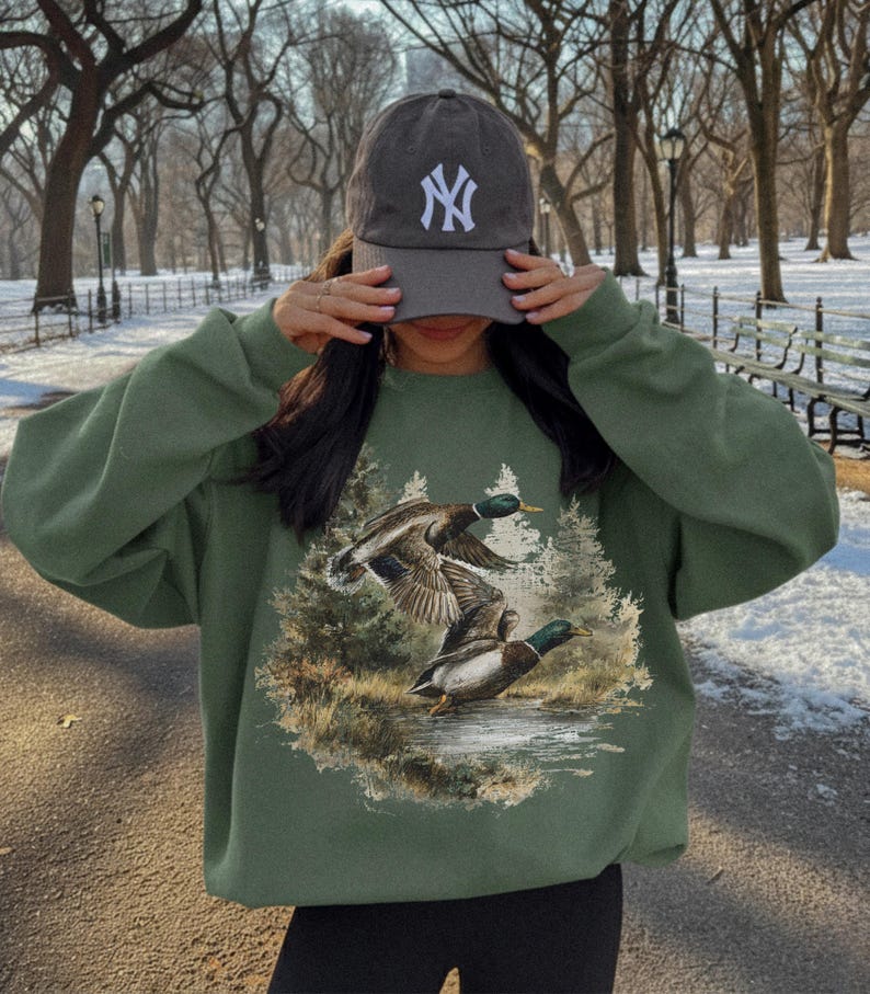 May include: An olive green sweatshirt featuring a graphic of two ducks in flight over a body of water, with a forest backdrop. The person is wearing a gray baseball cap with a white "NY" logo.