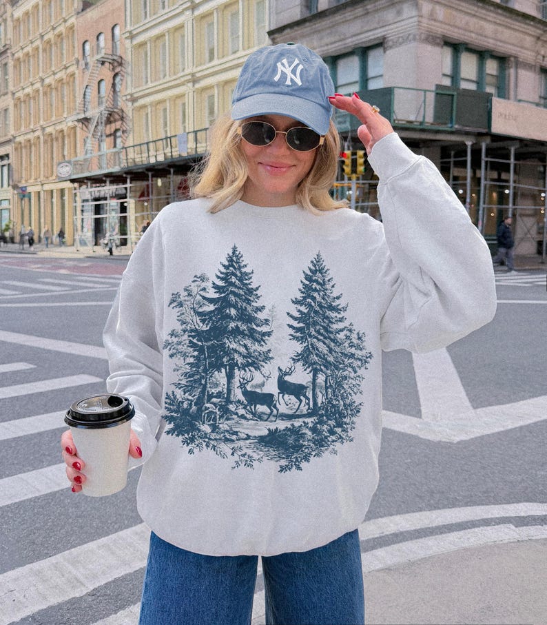 May include: A light gray sweatshirt featuring a forest scene with two deer and pine trees in a dark blue color. The person is wearing a light blue baseball cap with a white logo and sunglasses. They are holding a white coffee cup with a black lid.
