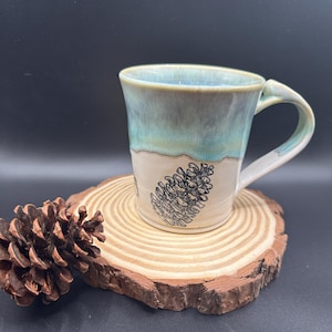May include: A handmade ceramic mug with a teal and beige glaze, featuring a black pinecone illustration. The mug sits on a wooden slice with a pinecone to the side. The mug has a curved handle.