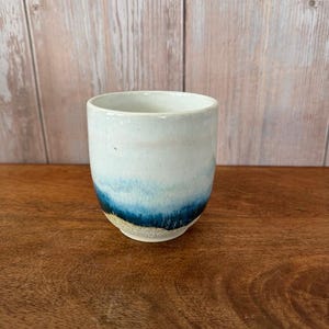 May include: A white ceramic cup with a blue and white glaze. The cup has a rounded shape and a slightly flared lip.