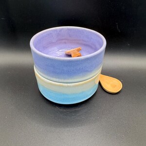 May include: A ceramic bowl with a light blue, white, and purple gradient design. A small wooden spoon rests on the side. Inside the bowl is a small, wooden, star-shaped object. The bowl is a handmade item.