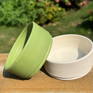 May include: Two ceramic bowls, one green and one white, are stacked on a wooden surface. The green bowl is on top of the white bowl.
