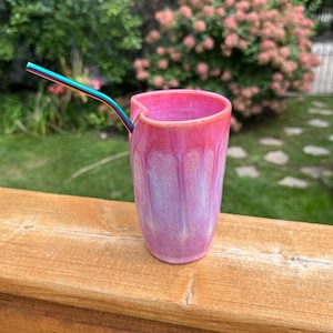 May include: A ceramic tumbler with a heart-shaped rim and a glossy gradient finish transitioning from deep pink at the top to a lighter lavender-pink at the base. A metallic rainbow-colored straw is inserted into the cup, extending outward.
