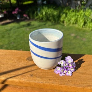 May include: A handmade ceramic cup with a cream-colored base and two diagonal blue stripes. The cup sits on a wooden surface next to a small cluster of purple flowers. The cup has a light blue rim.