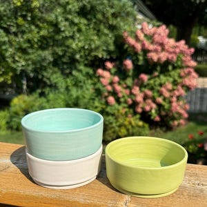 May include: Two ceramic bowls, one light green and one light yellow, are stacked on a wooden surface. The bowls are handmade and have a slightly textured finish.
