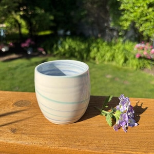 May include: A white ceramic tumbler with blue and green swirl design. The cup is on a wooden surface with a sprig of purple flowers. The background is a blurred view of green trees and grass.