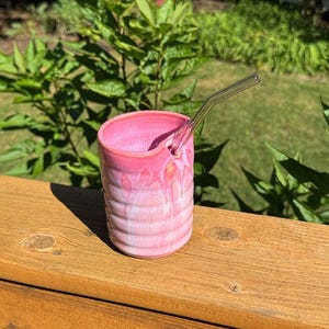May include: A cylindrical ceramic tumbler with a ribbed texture and a glossy pink gradient glaze. The cup features a specialized notch along the top rim designed to hold a clear, bent glass straw that extends upward from the opening.