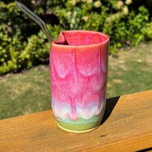 May include: A cylindrical ceramic tumbler featuring a vibrant gradient glaze that transitions from a deep pink at the top to a soft white and light green at the base. The rim has a distinct thumb indentation, and a clear glass straw is inserted.