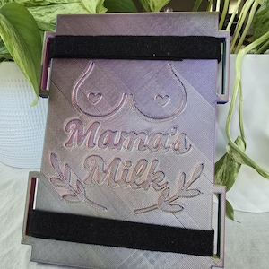 May include: A rectangular, iridescent breast milk storage container with the words "Mama's Milk" and a stylized illustration of breasts. The container has two black velcro straps.
