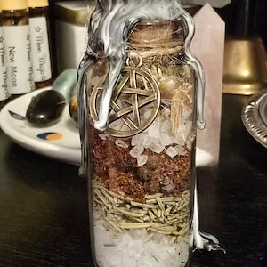 May include: A glass jar with a white and black wax seal, filled with layers of herbs, salt, and a pentagram charm. The jar is on a dark surface.