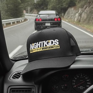 May include: A black trucker hat with the text "NIGHTKIDS" in white and yellow, sitting on the dashboard of a car. The car is driving on a road with another car in front. The background shows a forest.