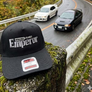 Op de afbeelding: Zwarte baseballcap met het woord "Emperor" in het wit geborduurd en Japanse tekst erboven. De cap ligt op een met mos bedekte stenen muur, met een kronkelende weg en auto's op de achtergrond.