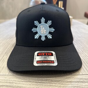 May include: Black baseball cap with a light blue embroidered design. The design features a sunburst with the letters "LA" in the center. The cap has a curved brim and a tag that reads "OTTO CLASSIC FIT OSFM MID PROFILE MAXIMUM COMFORT".