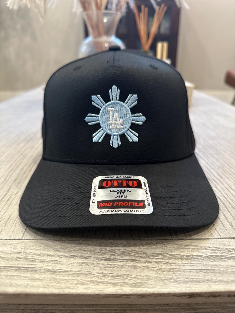 May include: Black baseball cap with a light blue embroidered design. The design features a sunburst with the letters "LA" in the center. The cap has a curved brim and a tag that reads "OTTO CLASSIC FIT OSFM MID PROFILE MAXIMUM COMFORT".