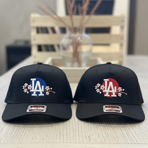 K&ouml;nnte beinhalten: Zwei schwarze Baseballkappen mit gesticktem "LA"-Schriftzug. Eine hat einen blauen Kreis hinter den Buchstaben, die andere einen roten Kreis. Beide Kappen sind mit floralen Designs verziert.