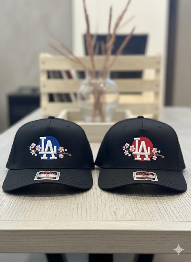 K&ouml;nnte beinhalten: Zwei schwarze Baseballkappen mit gesticktem "LA"-Schriftzug. Eine hat einen blauen Kreis hinter den Buchstaben, die andere einen roten Kreis. Beide Kappen sind mit floralen Designs verziert.