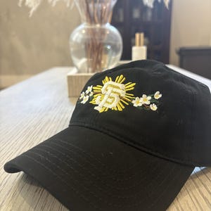May include: A black baseball cap featuring an embroidered design on the front. The embroidery displays a white and yellow stylized SF logo surrounded by a yellow sunburst and white flowers with green leaves on either side of the central emblem.