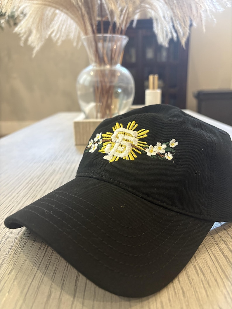 May include: A black baseball cap featuring an embroidered design on the front. The embroidery displays a white and yellow stylized SF logo surrounded by a yellow sunburst and white flowers with green leaves on either side of the central emblem.