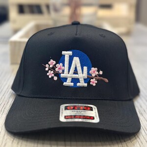 K&ouml;nnte beinhalten: Schwarze Baseballkappe mit den wei&szlig;en Buchstaben "LA" auf einem blauen Kreis, verziert mit rosa Kirschbl&uuml;tenstickerei. Die Kappe hat einen gebogenen Schirm und ein "Premium Fabric"-Etikett.
