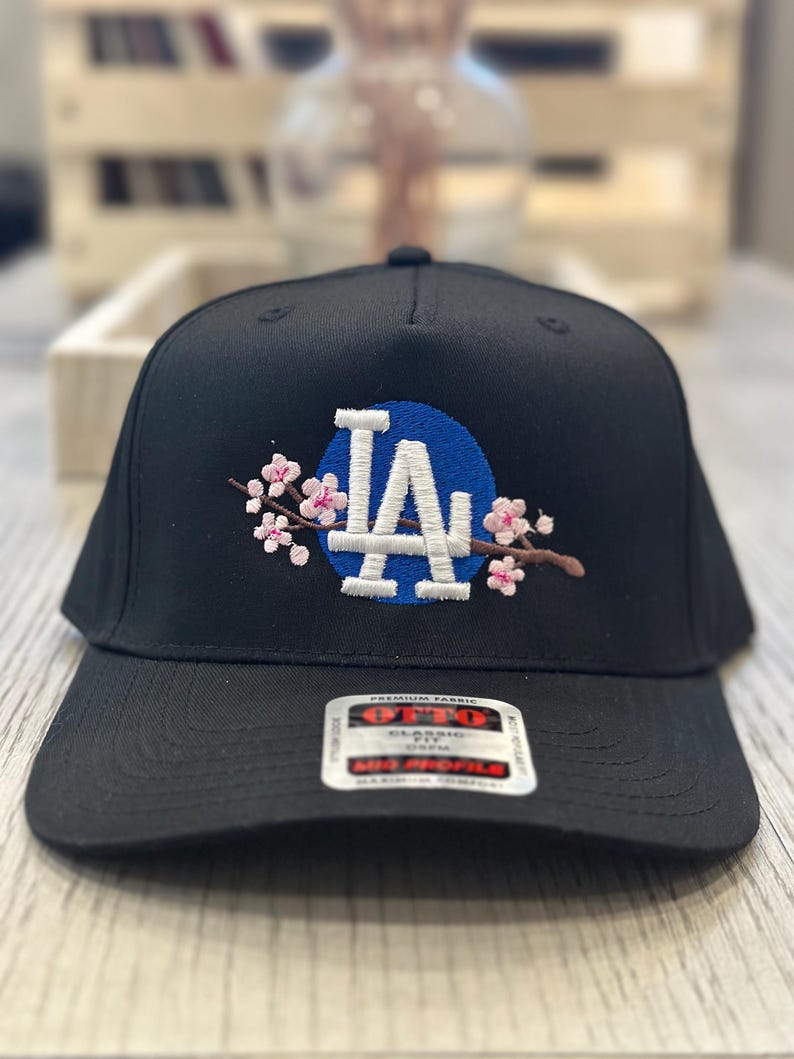 K&ouml;nnte beinhalten: Schwarze Baseballkappe mit den wei&szlig;en Buchstaben "LA" auf einem blauen Kreis, verziert mit rosa Kirschbl&uuml;tenstickerei. Die Kappe hat einen gebogenen Schirm und ein "Premium Fabric"-Etikett.