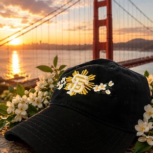 May include: A black baseball cap featuring an embroidered design on the front. The embroidery displays a yellow sunburst behind a white and yellow SF logo, flanked by small white flowers with green leaves on both sides of the central emblem.