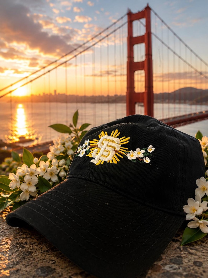 May include: A black baseball cap featuring an embroidered design on the front. The embroidery displays a yellow sunburst behind a white and yellow SF logo, flanked by small white flowers with green leaves on both sides of the central emblem.