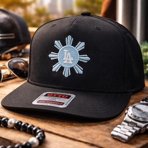 May include: Black baseball cap with a light blue and white embroidered design. The design features a sunburst with the letters "LA" in the center. The cap has a flat brim and a tag that reads "OTTO".