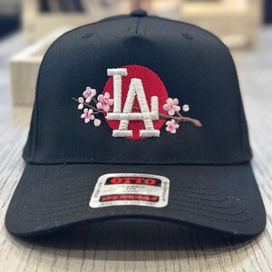 K&ouml;nnte beinhalten: Schwarze Baseballkappe mit den wei&szlig;en Buchstaben "LA" vor einem roten Kreis, verziert mit gestickten rosa Kirschbl&uuml;ten. Die Kappe hat einen gebogenen Schirm und ein Etikett mit der Aufschrift "OTTO".