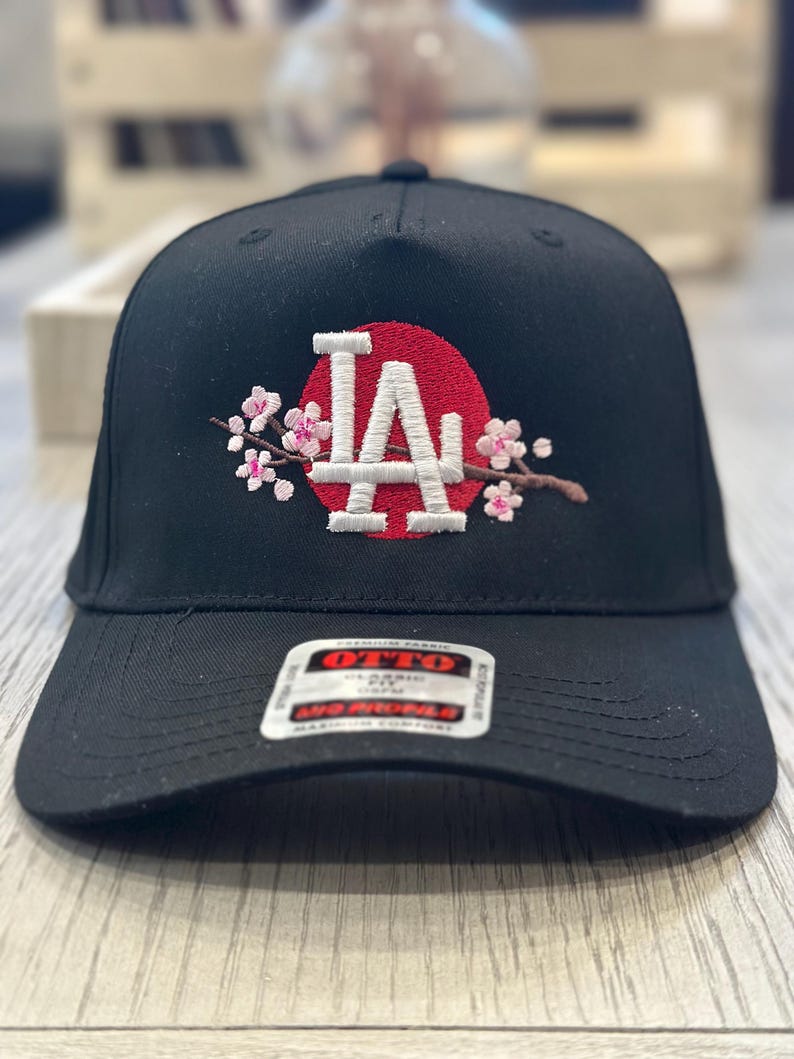 K&ouml;nnte beinhalten: Schwarze Baseballkappe mit den wei&szlig;en Buchstaben "LA" vor einem roten Kreis, verziert mit gestickten rosa Kirschbl&uuml;ten. Die Kappe hat einen gebogenen Schirm und ein Etikett mit der Aufschrift "OTTO".