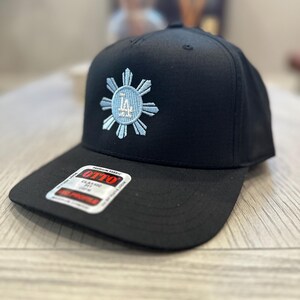 May include: Black baseball cap with a flat brim. The cap features a light blue and white embroidered design of a sunburst with the letters "LA" in the center. The cap has a "OTTO" label.