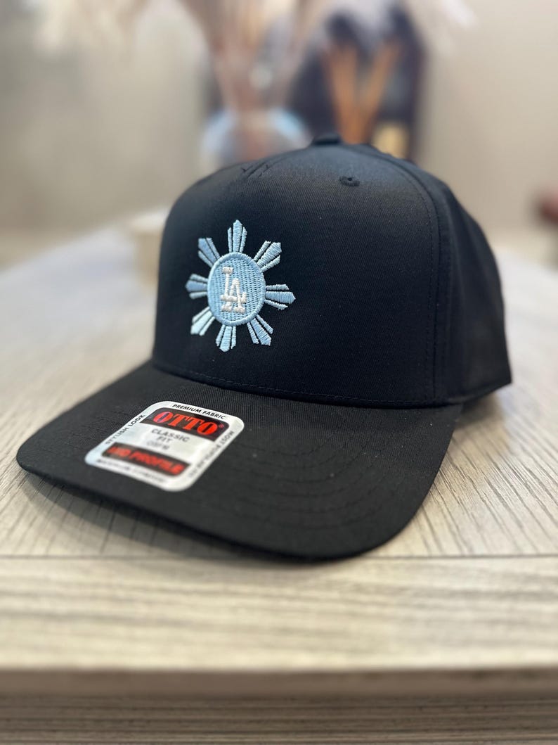 May include: Black baseball cap with a flat brim. The cap features a light blue and white embroidered design of a sunburst with the letters "LA" in the center. The cap has a "OTTO" label.