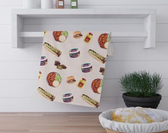 May include: A beige kitchen towel with a repeating watercolor pattern of various foods, including hot dogs, burgers, and containers of dip. The towel is hanging on a white shelf with jars and spices.