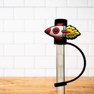 Può includere: Un tappo per bottiglia a forma di razzo nero e rosso con un motivo a fiamma gialla. Il tappo è attaccato a un cinturino in silicone nero.