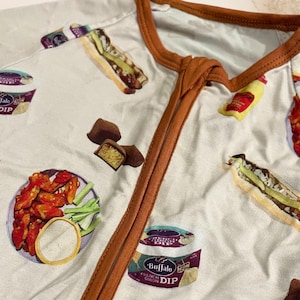 May include: White onesie with a repeating pattern of food items, including buffalo wings, sandwiches, and dip containers. The onesie has a brown trim around the collar and zipper. The overall design is a playful and colorful food-themed pattern.