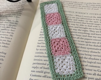 Könnte beinhalten: Ein rechteckiges Häkel-Lesezeichen mit einer vertikalen Reihe aus fünf abwechselnd weißen und hellrosa Granny Squares, umrandet von einer salbeigrünen Häkelkante. Das Lesezeichen liegt auf einer aufgeschlagenen Buchseite mit schwarzem Text.