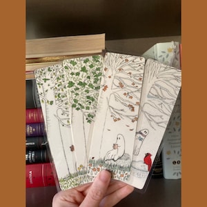 May include: A collection of whimsical bookmarks featuring illustrations of trees, ghosts, and autumn leaves. The bookmarks are held in a hand, with a bookshelf and books in the background. The bookmarks are in clear plastic sleeves.