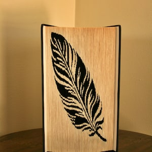 May include: A decorative book with a black feather design cut into the pages. The feather is a detailed silhouette against the light-colored pages. The book is standing upright on a wooden surface.
