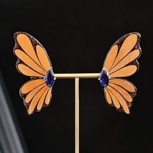 May include: A pair of butterfly wing earrings displayed on a gold-toned T-bar stand. Each earring features translucent orange wings with dark brown edges and a central marquise-cut deep blue gemstone, creating a symmetrical insect-inspired design.