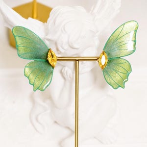 May include: A pair of earrings featuring translucent green butterfly wings with a subtle glitter finish, each accented by a single marquise-cut yellow crystal at the base, displayed on a thin gold-toned metal T-bar jewelry stand.