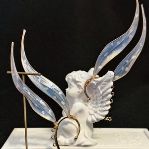 May include: A pair of gold-toned ear cuffs featuring translucent, iridescent wing-shaped attachments with embedded silver flakes. The cuffs have a curved wire frame with a series of small circular loops along the lower edge for additional adornment.