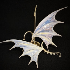 May include: An ear cuff featuring two iridescent, translucent dragon wings with a pearlescent finish and gold-toned wire detailing. The structure includes a curved wire frame with small circular loops and a gold-coloured metal display stand.