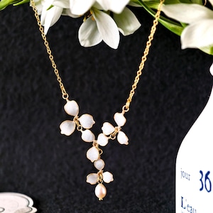 May include: A gold-coloured necklace with white, flower-shaped pendants and a single pearl drop. The necklace is set against a dark backdrop, with white flowers in the upper part of the image.