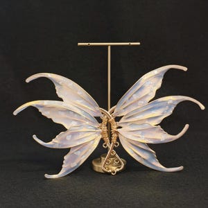 May include: A pair of iridescent, translucent wings with a delicate gold wire frame and embedded white flecks, mounted on a gold-toned metal T-shaped display stand. The wings feature curved, pointed edges and a soft, pearlescent colour gradient.