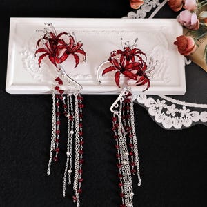 May include: A pair of elaborate earrings with crimson flower designs and silver detailing. Long, dangling chains with red and silver beads hang from the floral elements. The earrings are presented on a white rectangular surface.