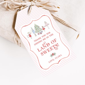 May include: Pink and white striped gift tag with a red border. The tag features a whimsical illustration of a Christmas tree, a nutcracker, and other characters. The text on the tag reads "Thank you for joining me in the Land of Sweets! Love, Clara".