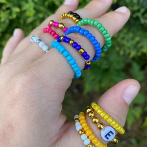May include: A set of colourful beaded rings, including a yellow ring with a white letter 'E' charm. The rings are stacked on a finger.
