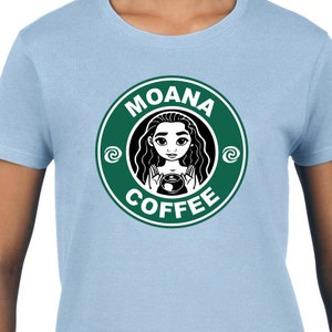 Puede incluir: Una camiseta azul claro con un logotipo circular verde que presenta una imagen de dibujos animados de Moana sosteniendo una taza de caf&eacute;. El texto del logotipo dice "Moana Coffee" con un dise&ntilde;o de espiral estilizado a cada lado del texto.