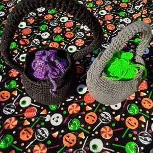 Op de afbeelding: Twee gehaakte Halloween-tassen. E&eacute;n is zwart met paars garen aan de binnenkant, de andere is grijs met groen garen aan de binnenkant. De tassen hebben handvatten en liggen op een zwarte stof met Halloween-snoep- en pompoenontwerpen.